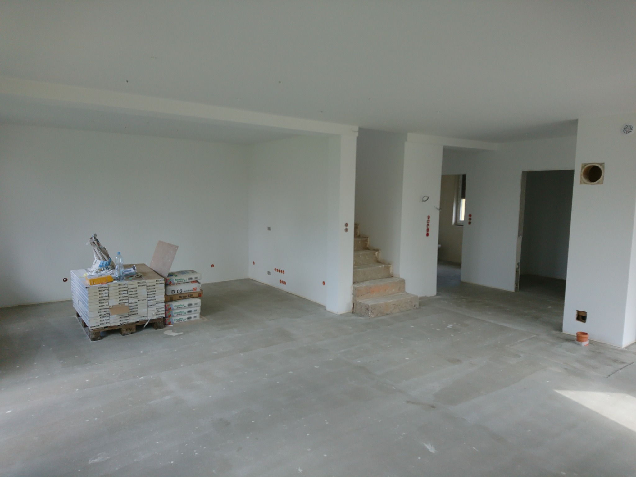 Surowy stan wnętrza domu w trakcie remontu: widoczne płyty karton-gips na palecie, surowe betonowe schody, ściany przygotowane do malowania z widocznymi puszkami elektrycznymi.