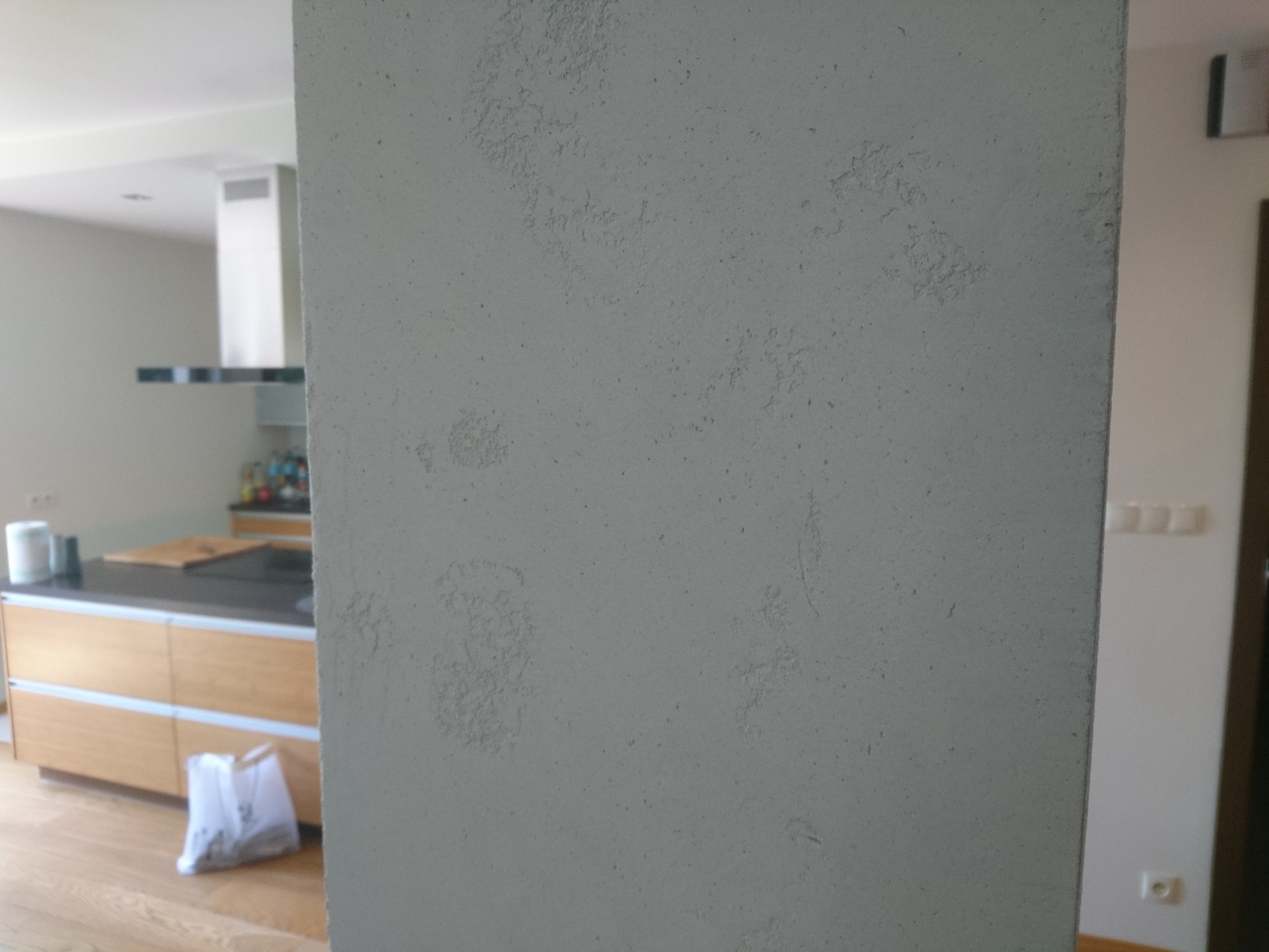 Szara ściana z dekoracyjną, nierówną fakturą na tle jasnej kuchni z drewnianymi szafkami i okapem.