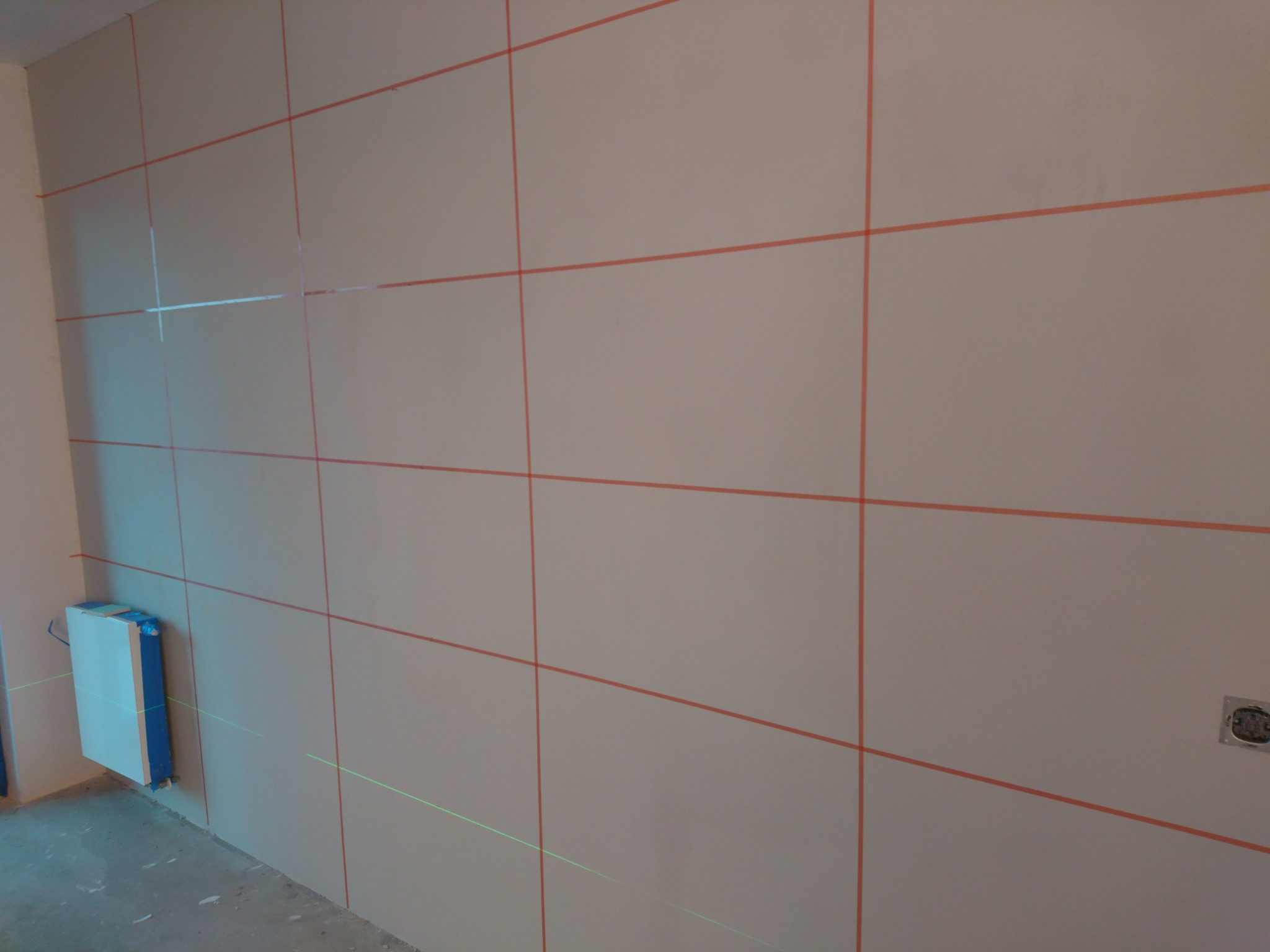 Ściana przygotowana do dalszych prac wykończeniowych, z widocznymi liniami poziomymi i pionowymi wyznaczonymi laserowo, obok ściany stoi urządzenie poziomujące.