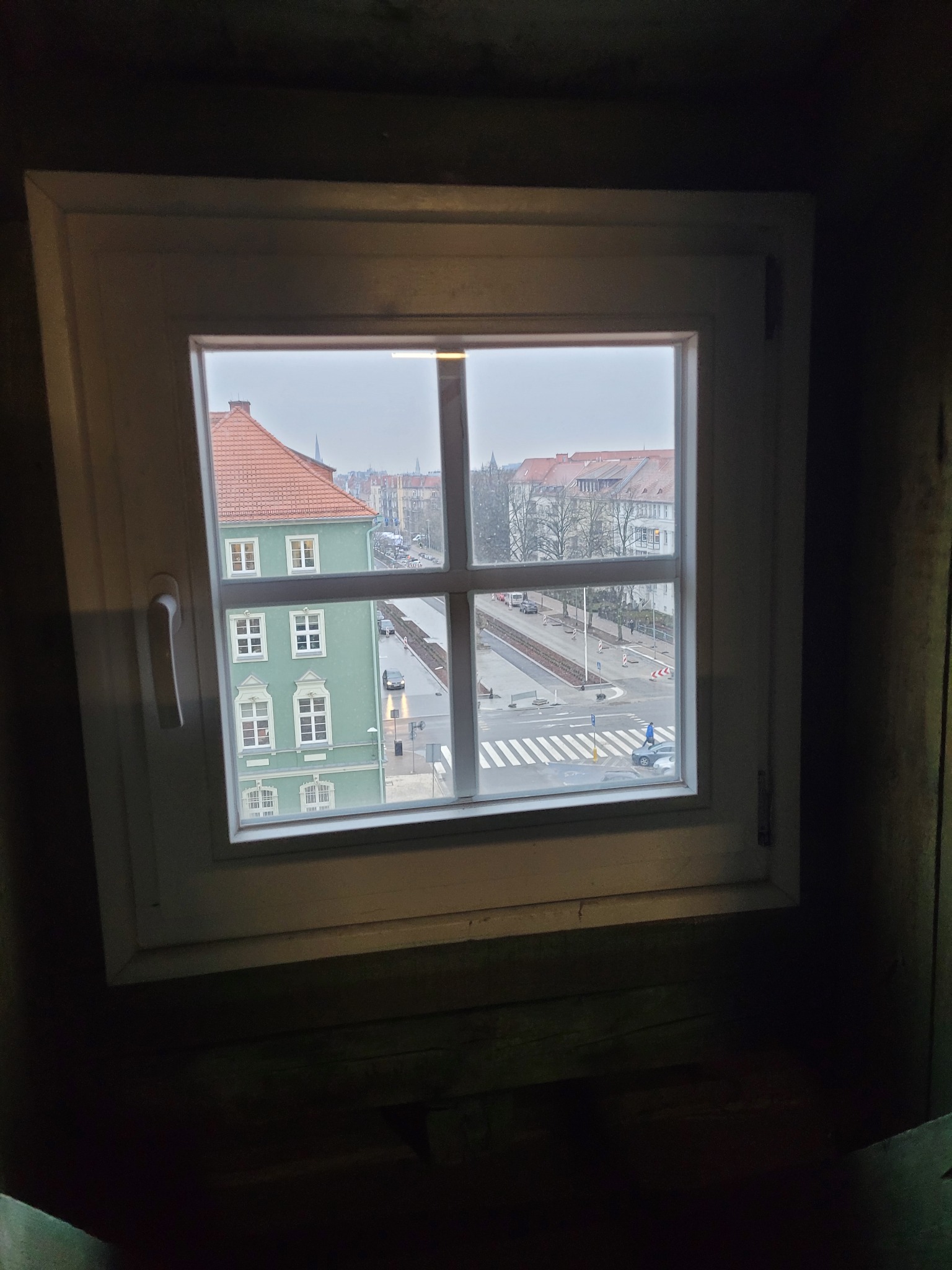 Widok z okna na ulicę miejską z przejściem dla pieszych i budynkami w tle, okno z białymi ramami osadzone w ciemnym, drewnianym wnętrzu.