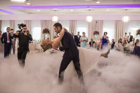 Para młoda tańczy taniec weselny w dymie, nagrywana przez kamerzystę na tle gości weselnych w sali bankietowej.