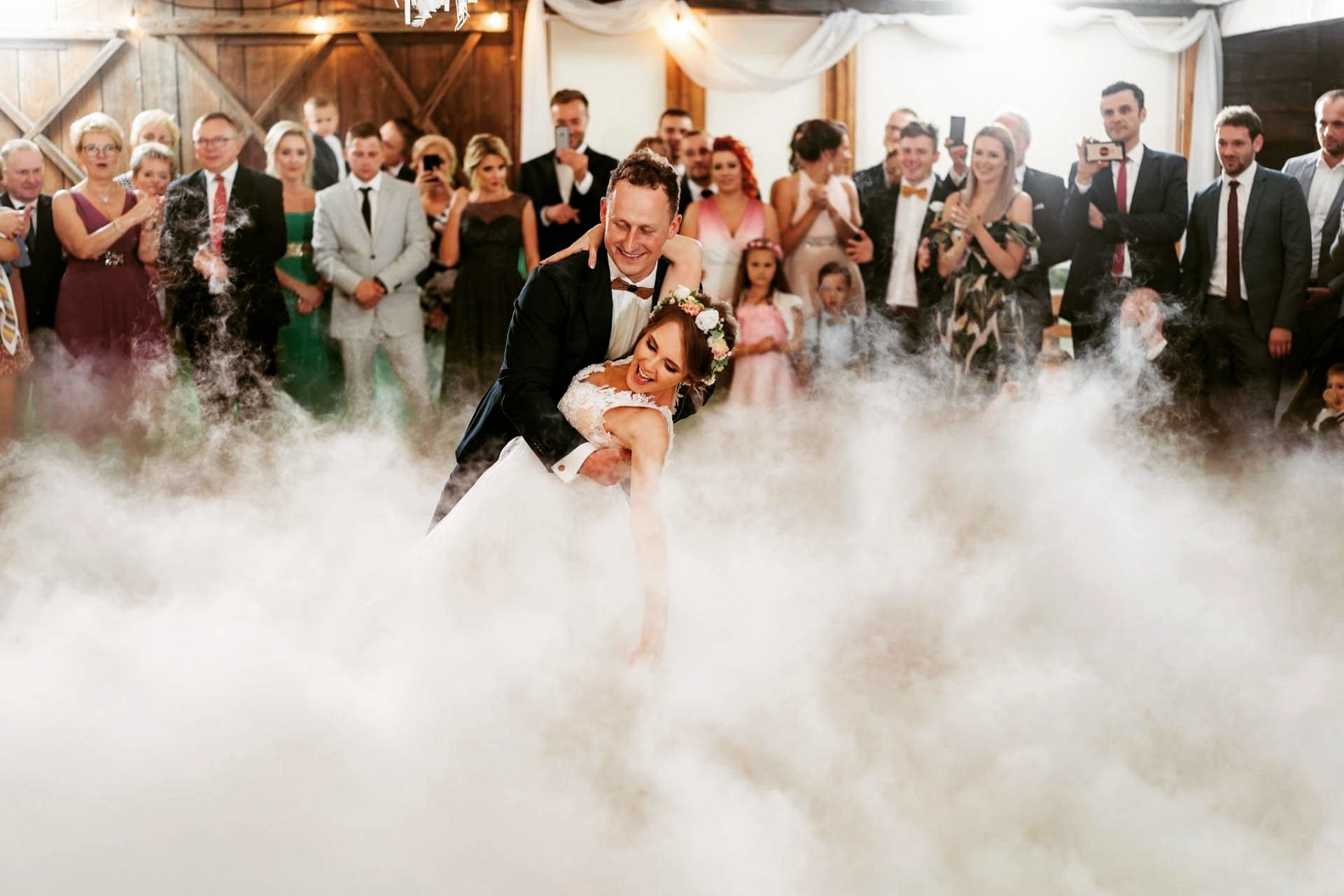 Para młoda w pierwszym tańcu, unosząca się w gęstej mgle na tle gości weselnych w sali rustykalnej z drewnianymi elementami.