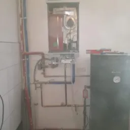 Zamontowany kocioł grzewczy Kospel z podłączonymi rurami miedzianymi i plastikowymi, obok zbiornik na wodę, poziomica na górze kotła, białe płytki na ścianie.