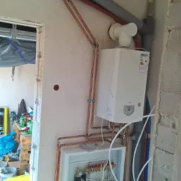 Montaż instalacji grzewczej w surowym wnętrzu: miedziane rury, biały piec gazowy z etykietą energetyczną, rozdzielacz w białej obudowie, szare rury wentylacyjne i instalacyjne, widoczne...