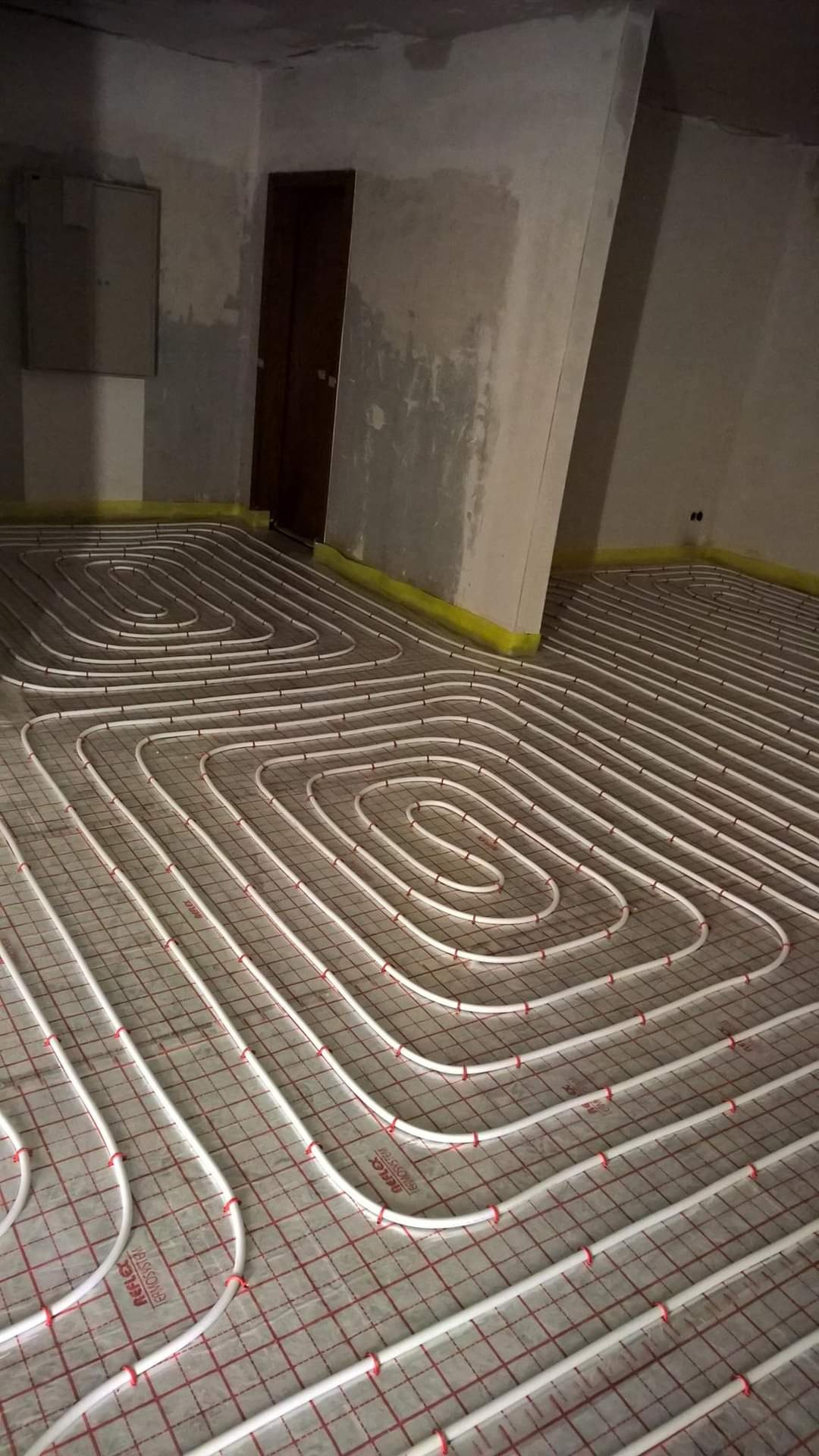 Białe rury systemu ogrzewania podłogowego ułożone na siatce w surowym pomieszczeniu z betonowymi ścianami i żółtą taśmą przy podłodze.