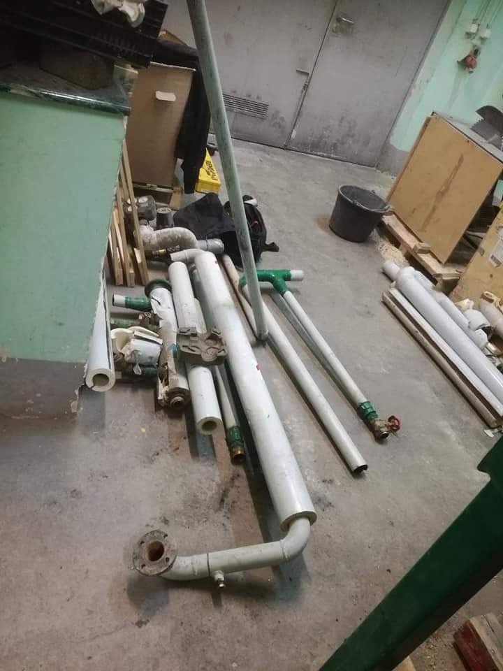 Stos rur PCV i metalowych złączek, niektóre z zaworami, leżące na betonowej podłodze w magazynie lub warsztacie, obok wiaderka i kartonów.