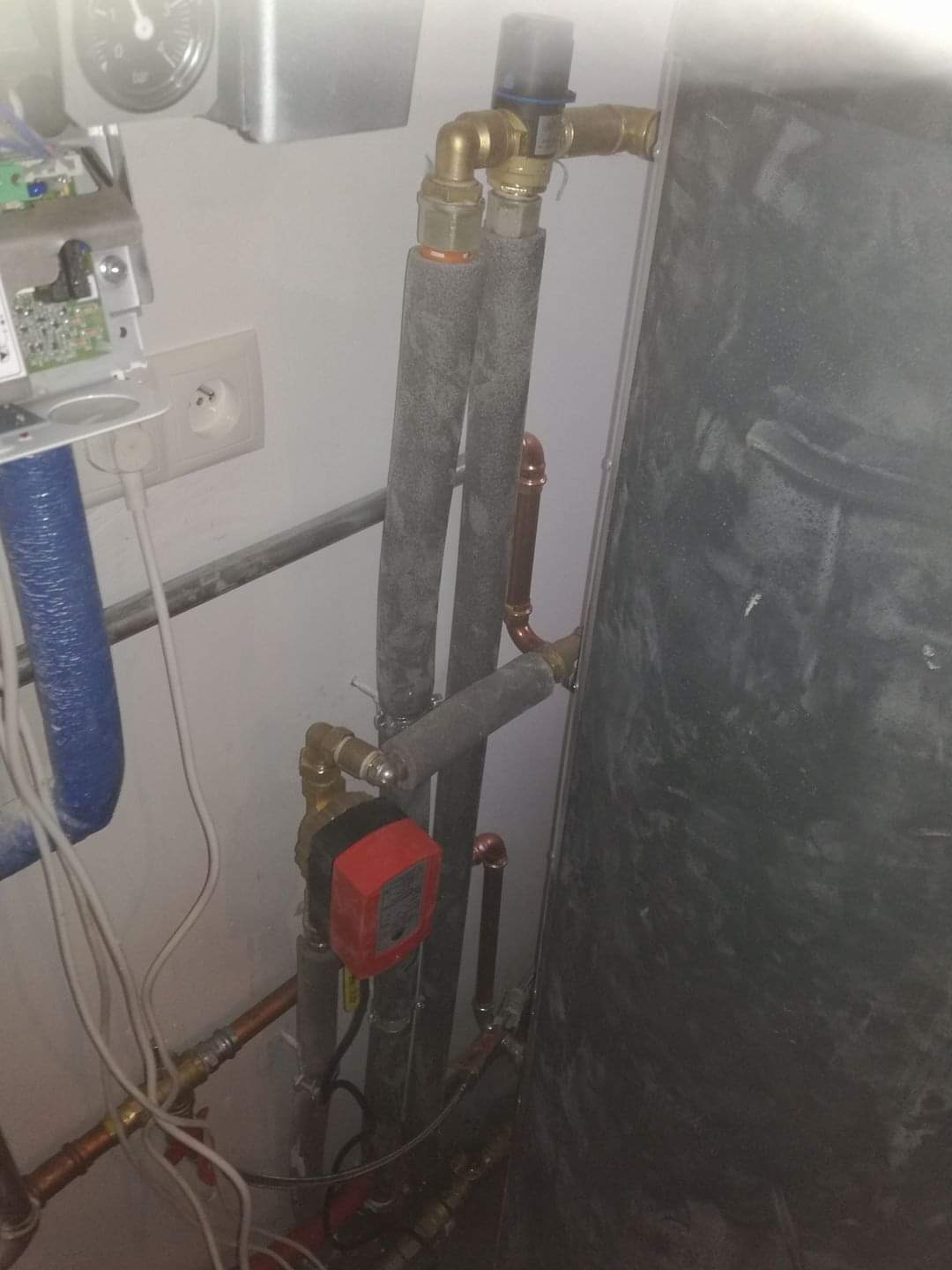 Instalacja centralnego ogrzewania z widocznym zbiornikiem, pompą obiegową z czerwoną obudową i zaizolowanymi rurami miedzianymi w kotłowni. Manometr na ścianie.