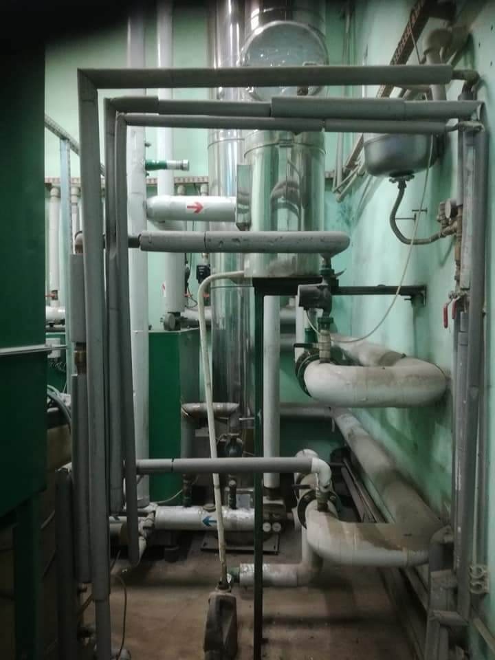 Złożony system rur w kotłowni z metalowym zbiornikiem, widoczne zawory, manometry i izolacja termiczna na rurach, ściany w odcieniach zieleni.