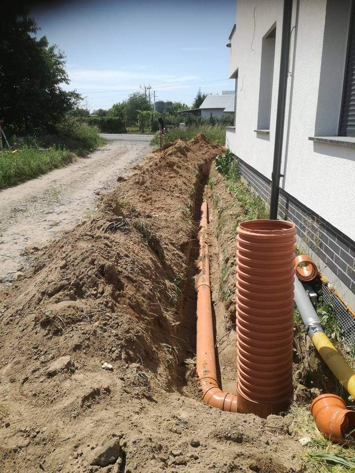 Pomarańczowe rury kanalizacyjne ułożone w wykopie wzdłuż ściany budynku, z widoczną studzienką i żółtą rurą wentylacyjną, na tle ziemi i zieleni.