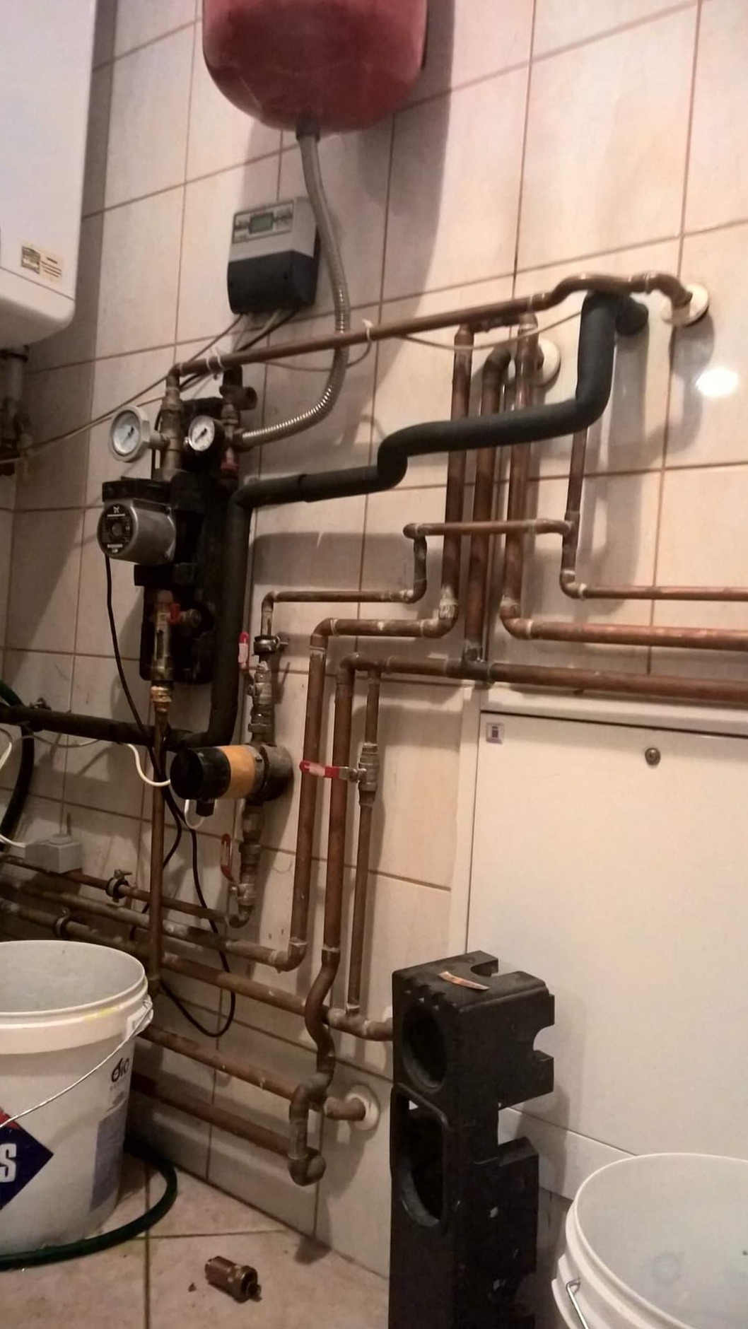 Złożona instalacja grzewcza z kotłem, zbiornikiem wyrównawczym, pompami obiegowymi i rurami miedzianymi na tle kafelkowanej ściany, widoczny odkręcony zawór i wiadro na podłodze.
