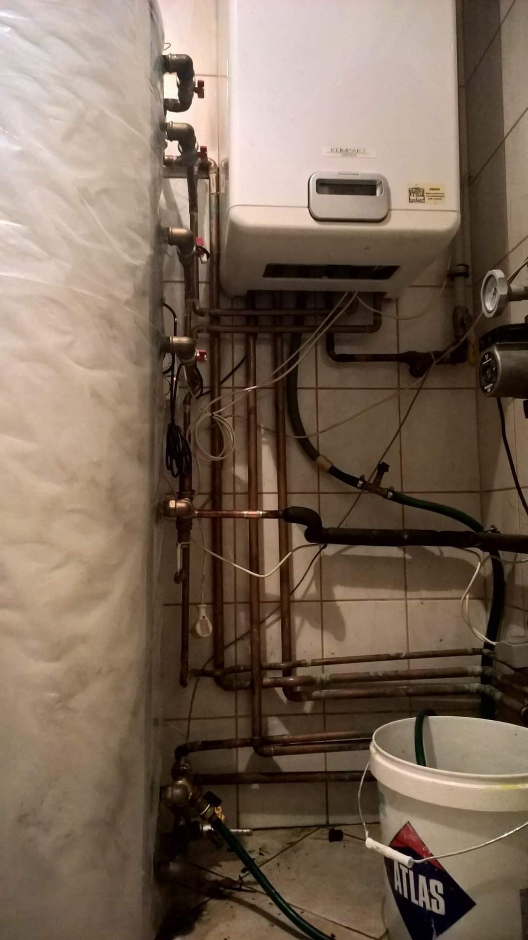Kompaktowy piec gazowy wiszący na ścianie z widocznymi rurami miedzianymi, zaworami i pompą obiegową, przy ścianie osłonięty zbiornik na wodę, białe kafelki na ścianie i wiadro z napisem ATLAS.