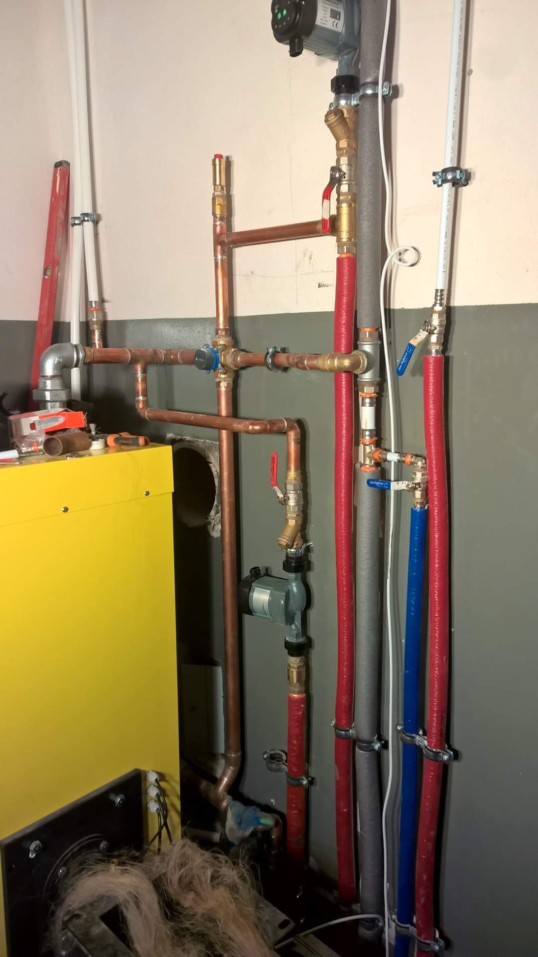 Miedziane i plastikowe rury instalacji wodnej i centralnego ogrzewania z zaworami i pompą obiegową zamocowane do ściany, widoczny fragment żółtego kotła grzewczego i narzędzia.