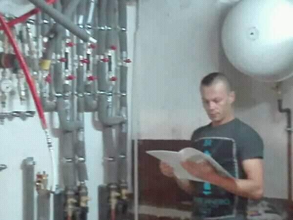 Instalator przegląda dokumentację techniczną na tle rozbudowanej instalacji grzewczej z licznymi zaworami i przewodami, obok znajduje się duży zbiornik wyrównawczy.