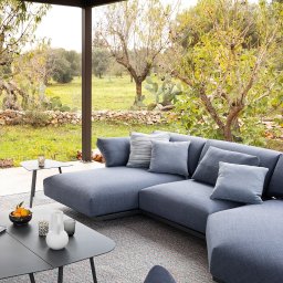 Tarcon Tarasy - Zadaszony taras z nowoczesnym, narożnym, szarym zestawem wypoczynkowym, stolikami kawowymi, ceramiką dekoracyjną i widokiem na ogród z kaktusami.