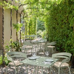 Tarcon Tarasy - Zacieniona przestrzeń z metalową pergolą porośniętą zielenią, pod którą stoją stoliki z nakryciem i krzesłami w kolorze pistacjowym, całość na żwirowym podłożu.