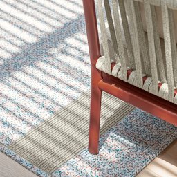 Tarcon Tarasy - Fragment krzesła z beżową plecionką i czerwonym stelażem na wzorzystym dywanie w odcieniach bieli, błękitu i czerwieni, oświetlonym promieniami słońca padającymi przez żaluzje.