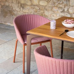 Tarcon Tarasy - Zestaw mebli ogrodowych: drewniany stół z metalowymi nogami, dwa różowe krzesła z tapicerowanymi siedziskami, naczynia i dekoracyjny lampion na tle kamiennej ściany.
