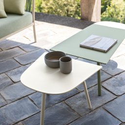 Tarcon Tarasy - Dwa stoliki kawowe na zewnątrz, jeden kremowy z miseczką i kubkiem, drugi zielony z książką 'Sensual Living', na posadzce z kamiennych płyt, w tle zieleń drzew.