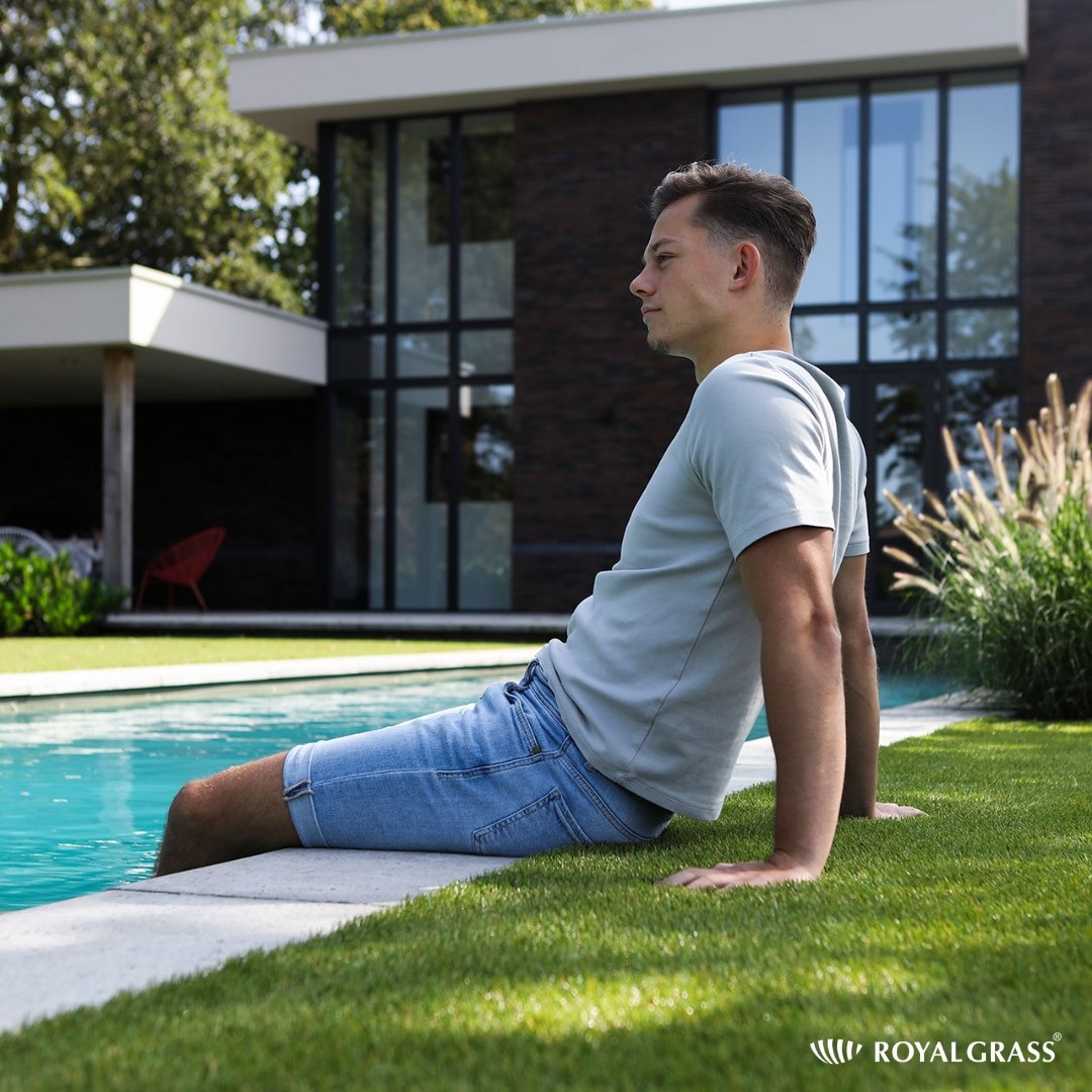 Młody mężczyzna siedzi na sztucznej trawie przy basenie, nowoczesny dom z ciemnej cegły w tle, dekoracyjne trawy ozdobne obok basenu.