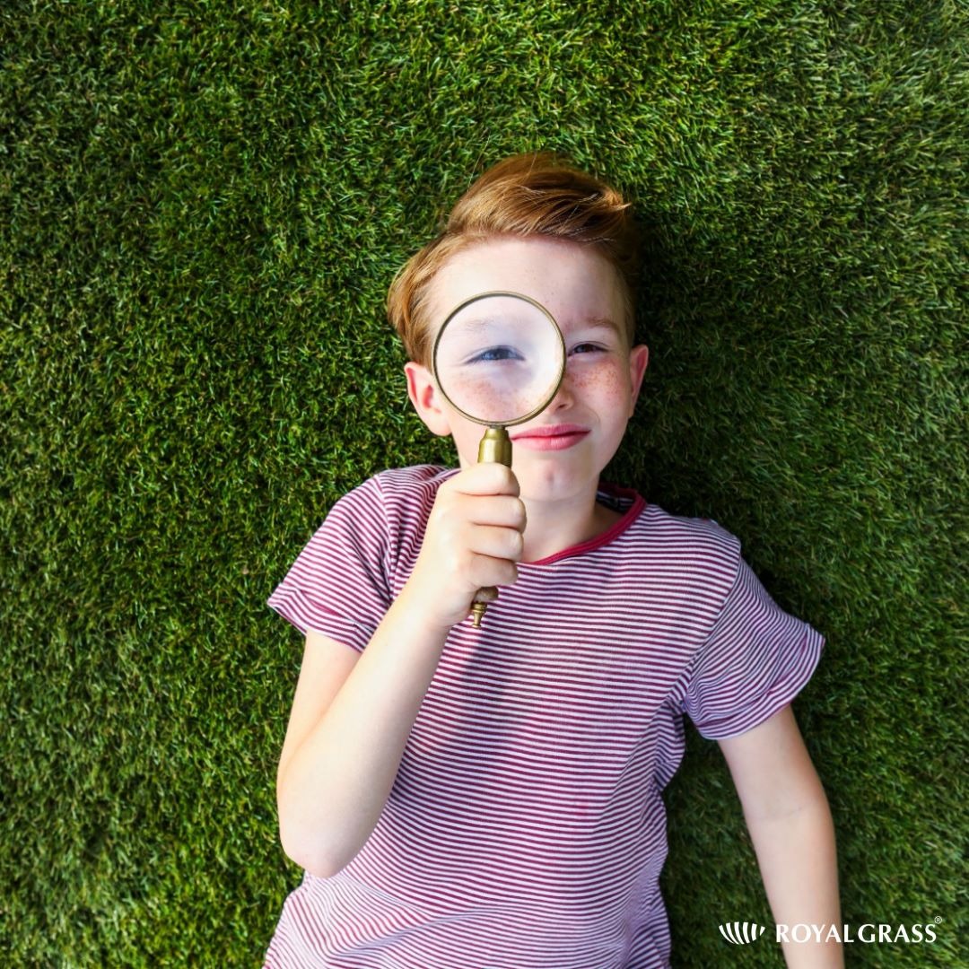 Chłopiec z rudymi włosami leży na intensywnie zielonej trawie, przyglądając się jej przez lupę z mosiężną ramką, uśmiechając się do kamery.