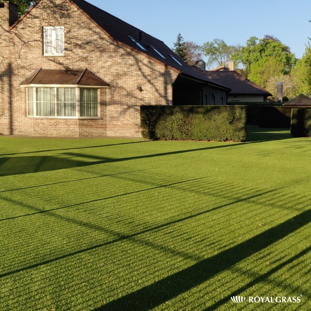 Rozległy trawnik z syntetycznej trawy przed ceglanym domem z oknem wykuszowym, cień rzucany przez konstrukcję, logo Royal Grass w prawym dolnym rogu.