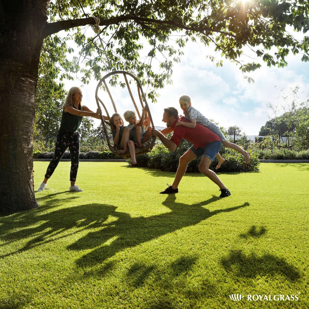 Rodzina bawi się na trawniku z syntetycznej trawy, dwoje dzieci na wiklinowej huśtawce pod drzewem, ojciec z dzieckiem na barana, jasne słońce i cień drzewa na zielonej trawie.