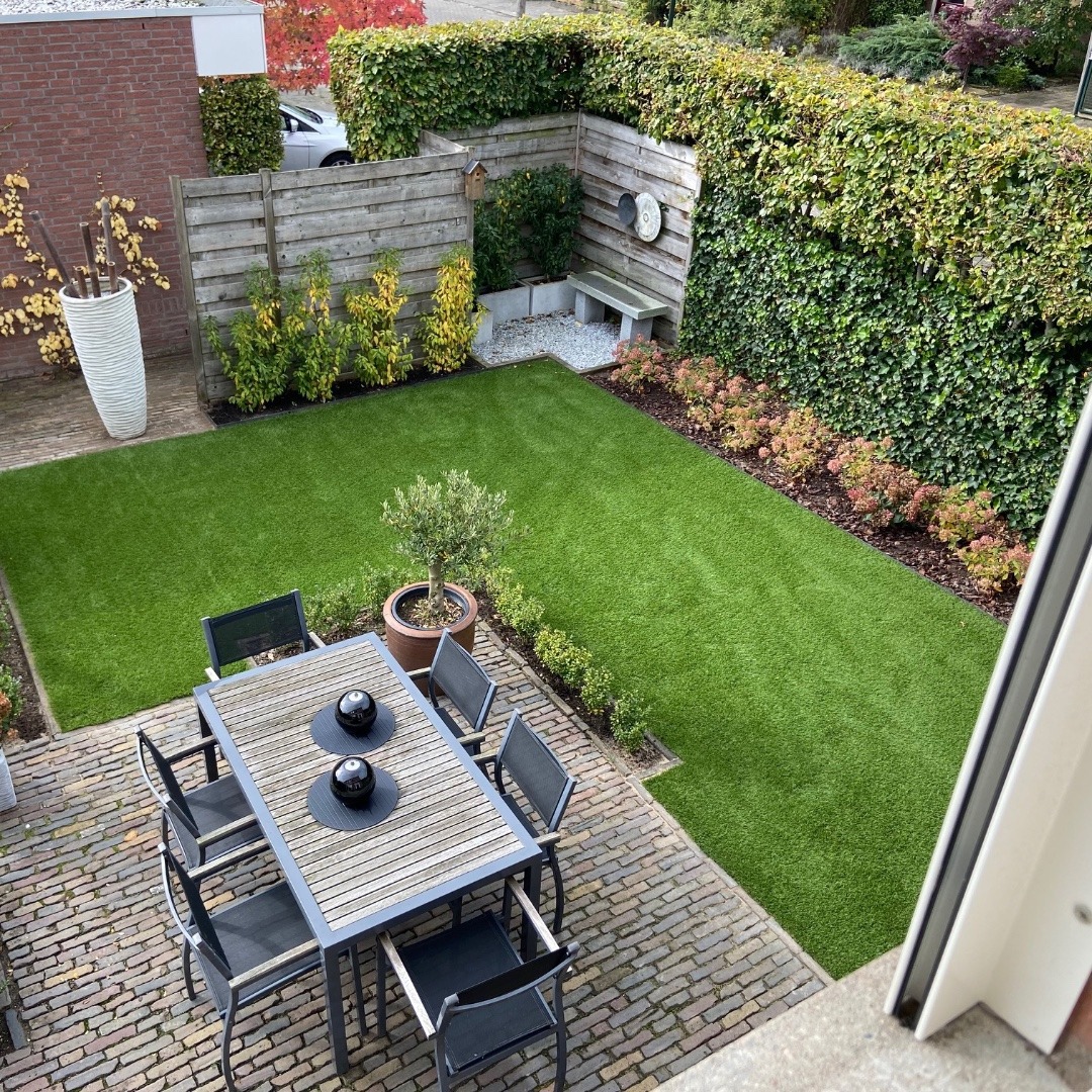 Widok z góry na zadbany ogród z równo przystrzyżonym trawnikiem, drewnianym stołem z czarnymi dekoracjami, żywopłotem i elementami małej architektury ogrodowej.