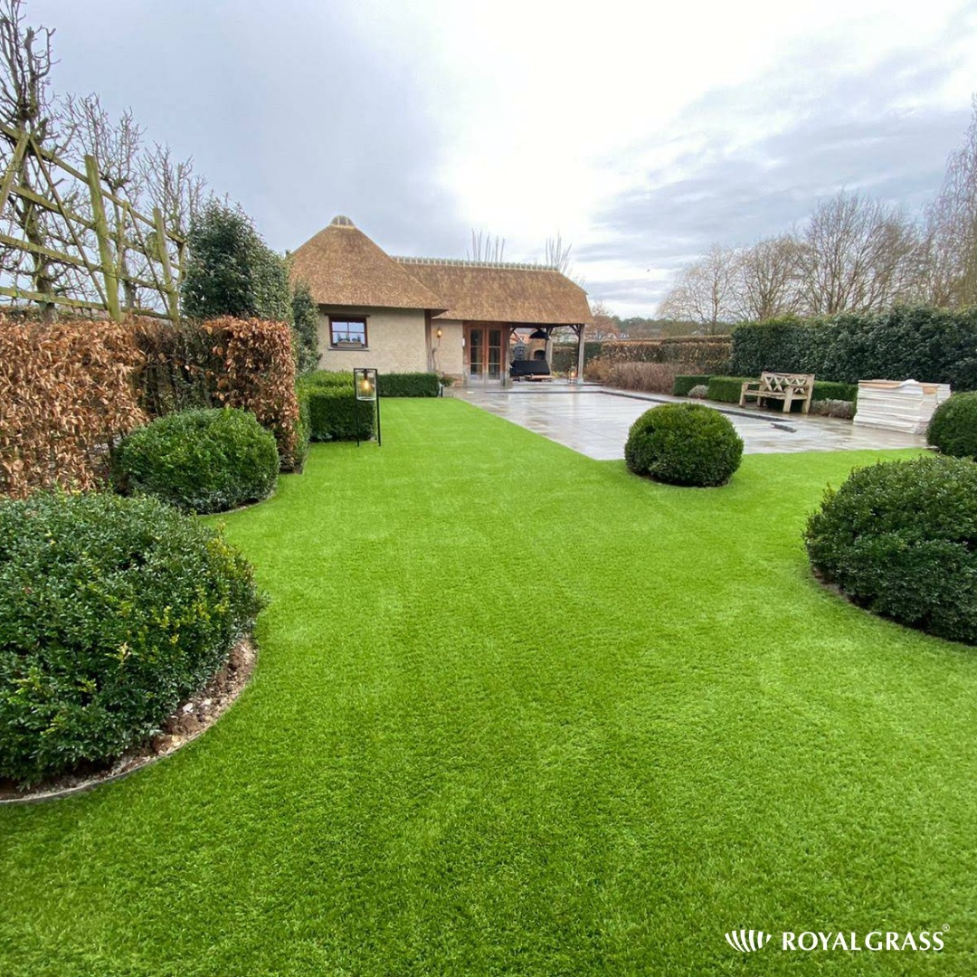 Ogród z intensywnie zieloną, sztuczną trawą, okrągłymi krzewami, brukowanym tarasem i domem krytym strzechą w tle.