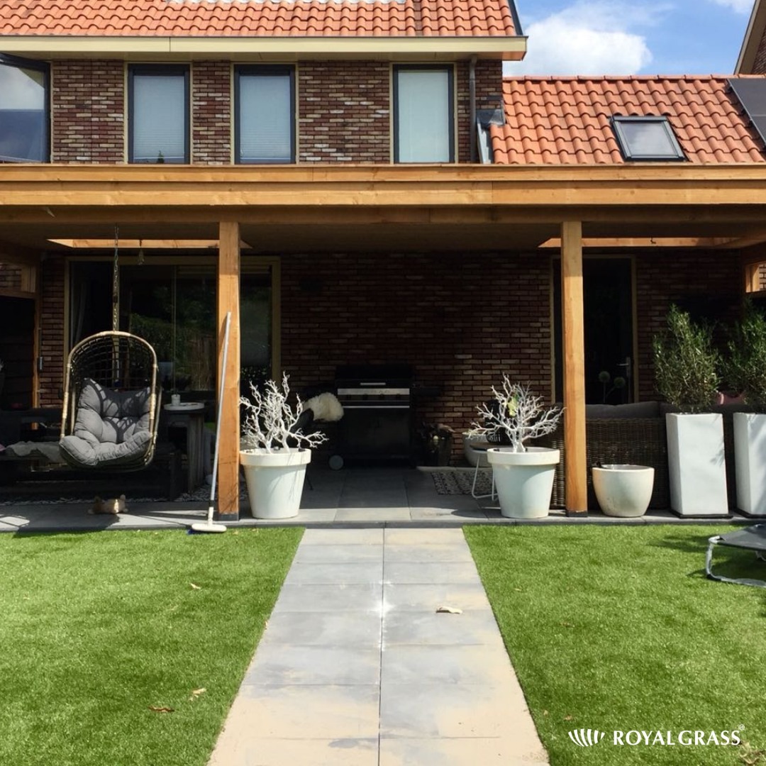 Zadaszony taras z cegły z drewnianą konstrukcją, sztuczna trawa i ścieżka z płyt, wiszące krzesło i dekoracyjne rośliny w donicach.