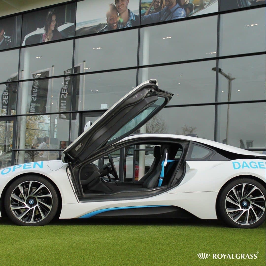 Biały samochód sportowy z otwartymi drzwiami typu skrzydła mewy, zaparkowany na sztucznej trawie przed szklanym budynkiem z fotografiami osób.