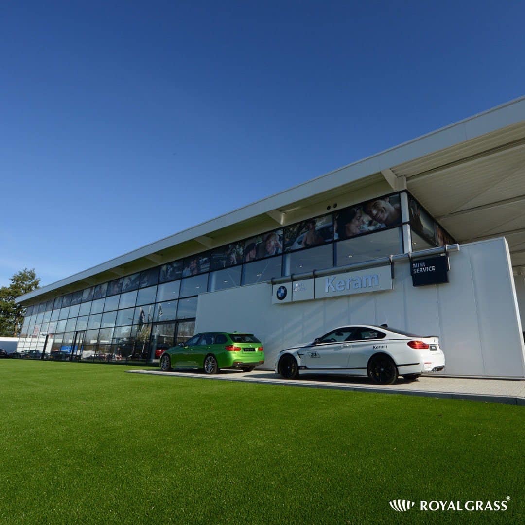 Salon samochodowy z dużym oknem i sztuczną trawą przed budynkiem, prezentacja dwóch samochodów.