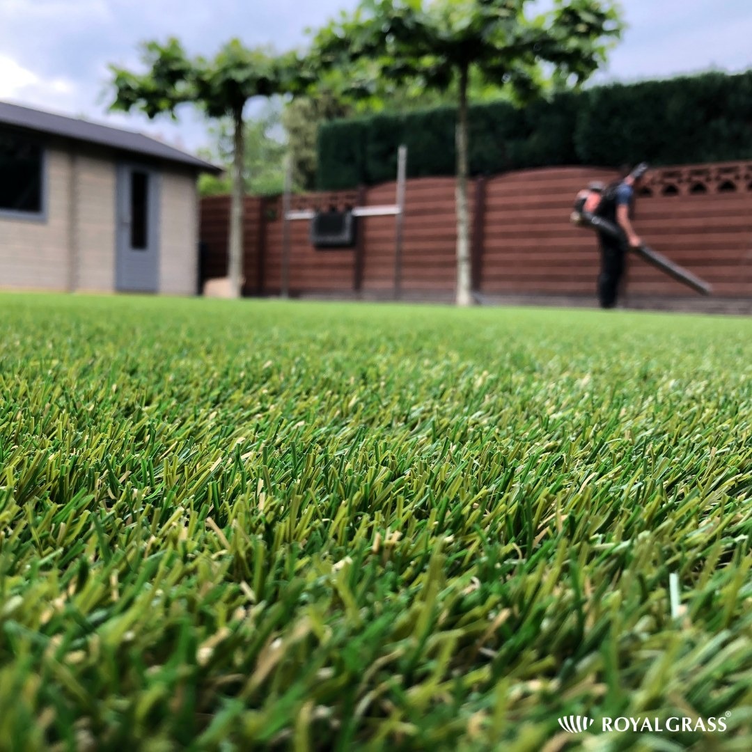 Niski kąt widzenia na zielony trawnik z trawy syntetycznej, w tle domek narzędziowy, drzewa formowane i pracownik z dmuchawą do liści przy brązowym ogrodzeniu.