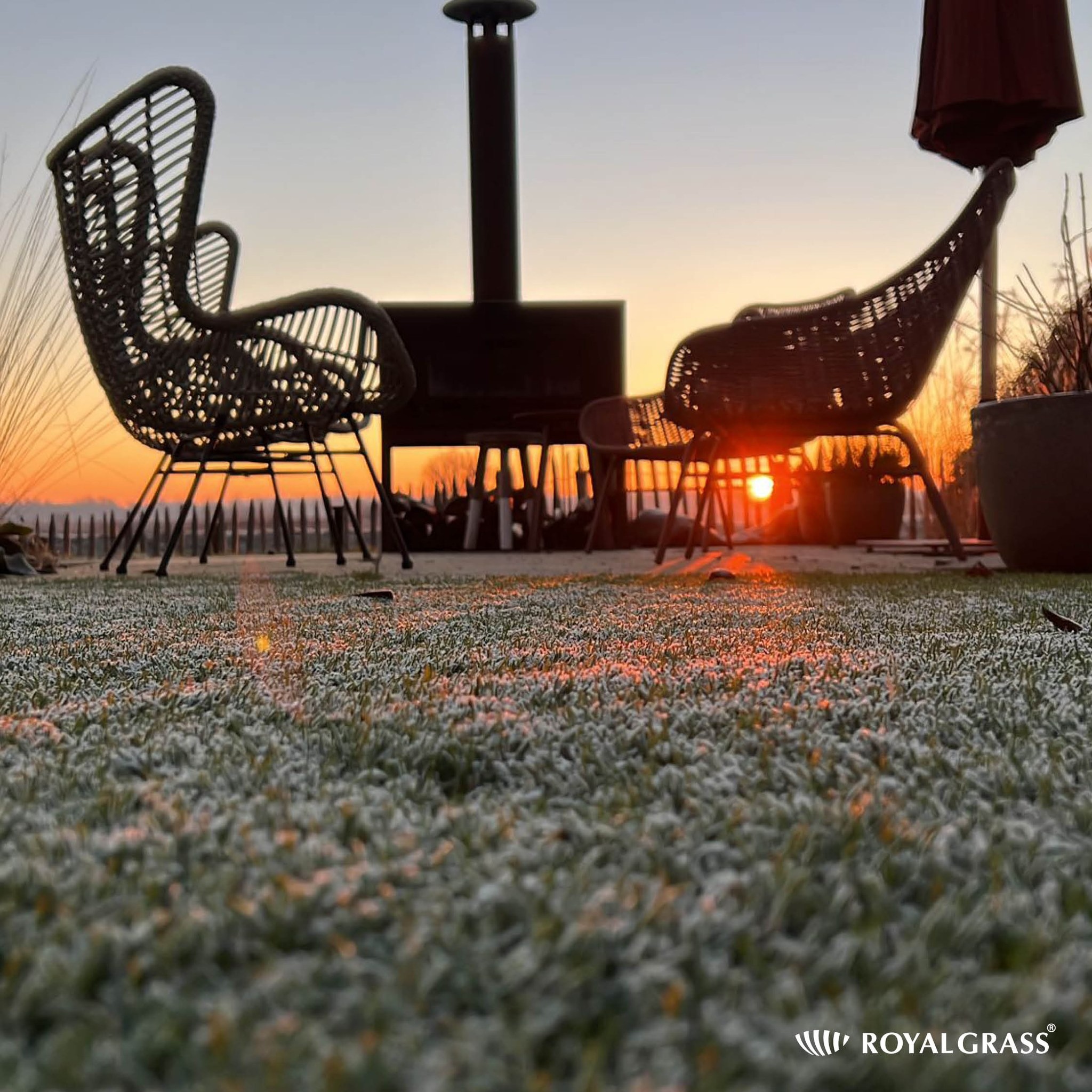 Dwa wiklinowe fotele i palenisko na tarasie z sztuczną trawą pokrytą szronem, w tle wschodzące słońce.