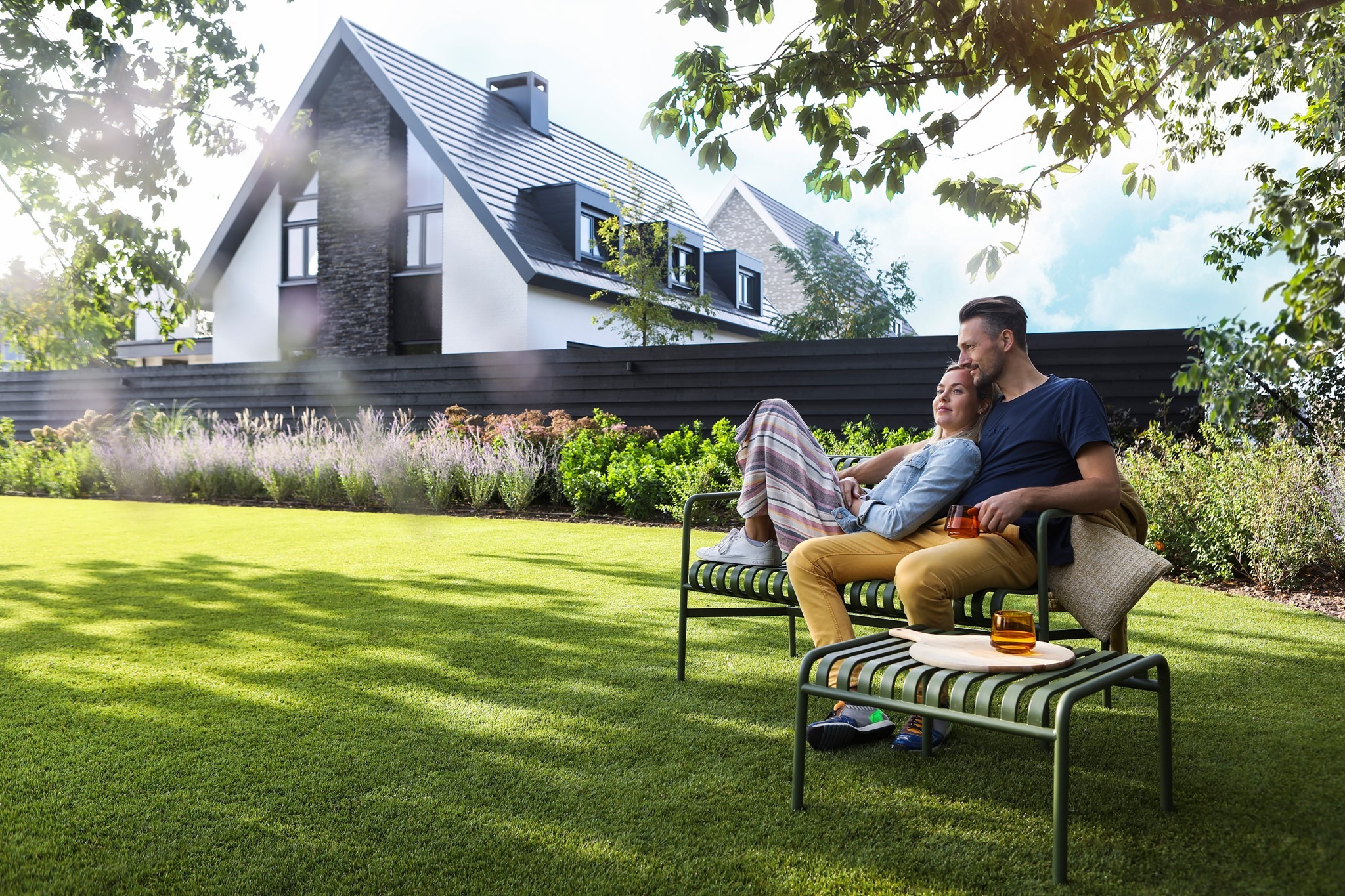 Para relaksuje się na nowoczesnym metalowym zestawie ogrodowym na idealnie przystrzyżonym trawniku; w tle nowoczesny dom z czarnym dachem i elementami z kamienia.