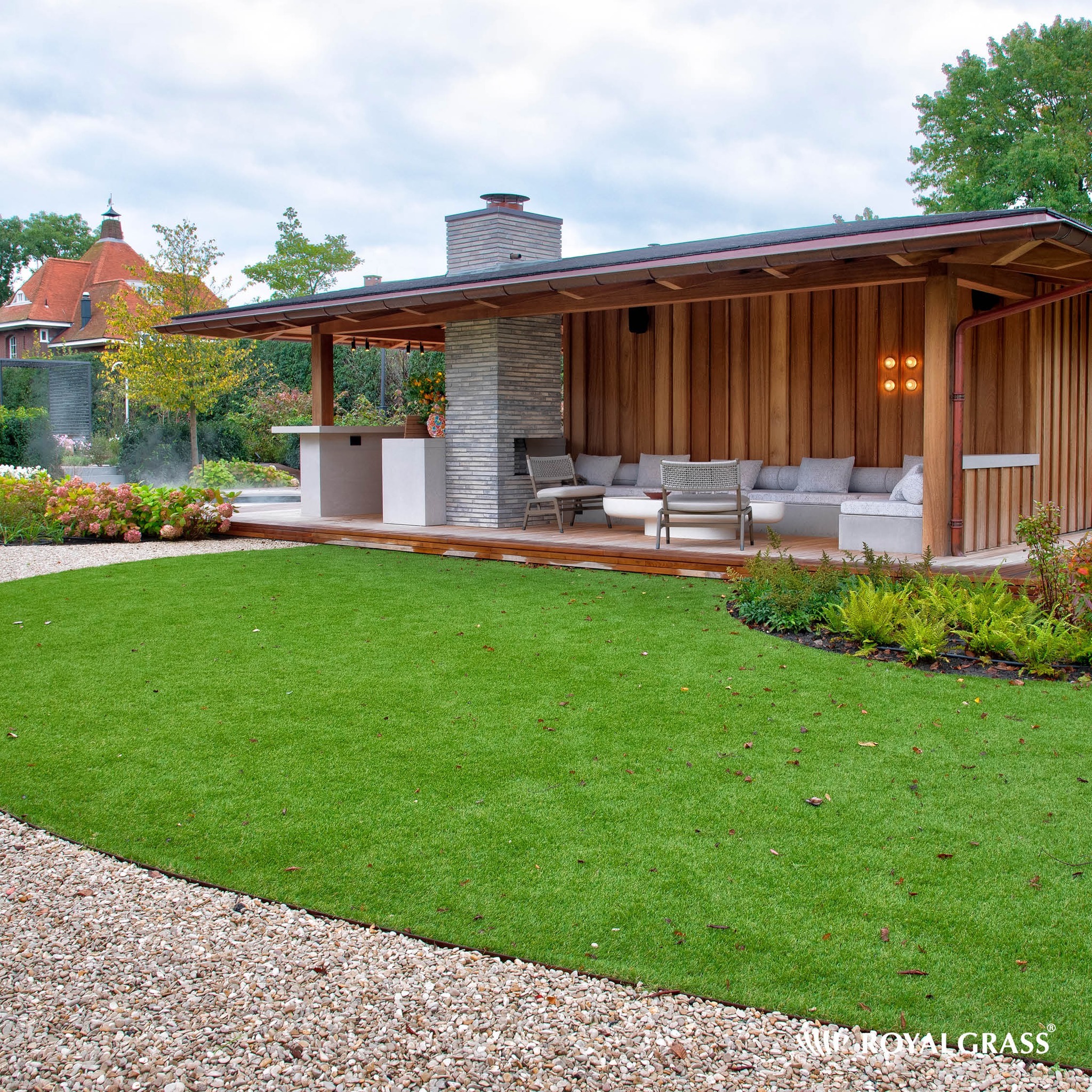 Nowoczesny ogród z drewnianą altaną, zewnętrznym kominkiem z szarej cegły, sztuczną trawą i żwirową ścieżką.