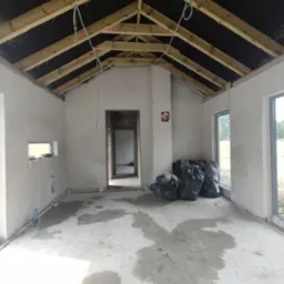 Wnętrze w stanie surowym otwartym: ściany po tynkowaniu, widoczne instalacje, drewniana konstrukcja dachu, okna z widokiem na zewnątrz i worki z odpadami budowlanymi.