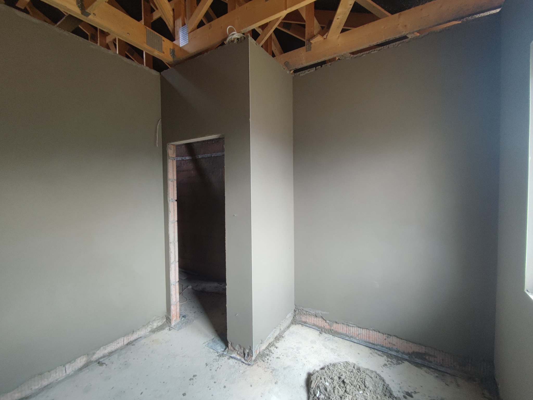 Świeżo nałożony, gładki tynk maszynowy na ścianach wewnętrznych pomieszczenia w stanie surowym, widoczne drewniane elementy konstrukcji dachu i fragment otworu drzwiowego.