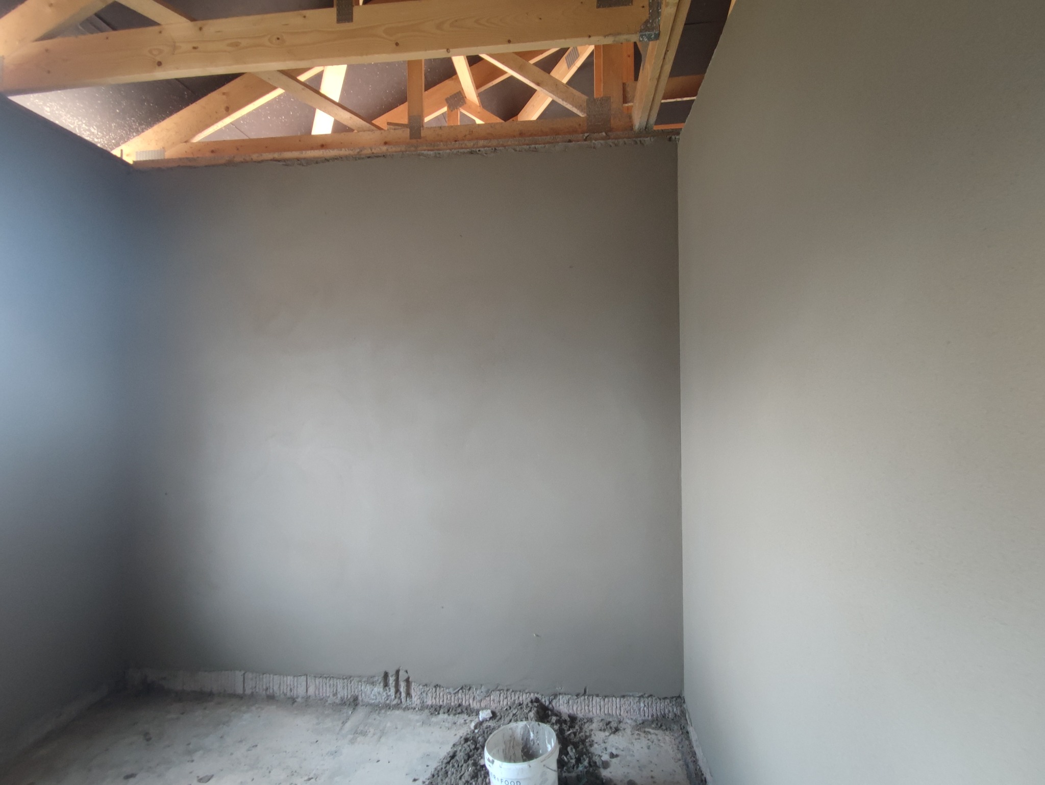 Świeżo nałożona, gładka, szara warstwa na ścianach w trakcie prac wykończeniowych, widoczna konstrukcja drewniana dachu i wiadro z resztkami zaprawy na podłodze.