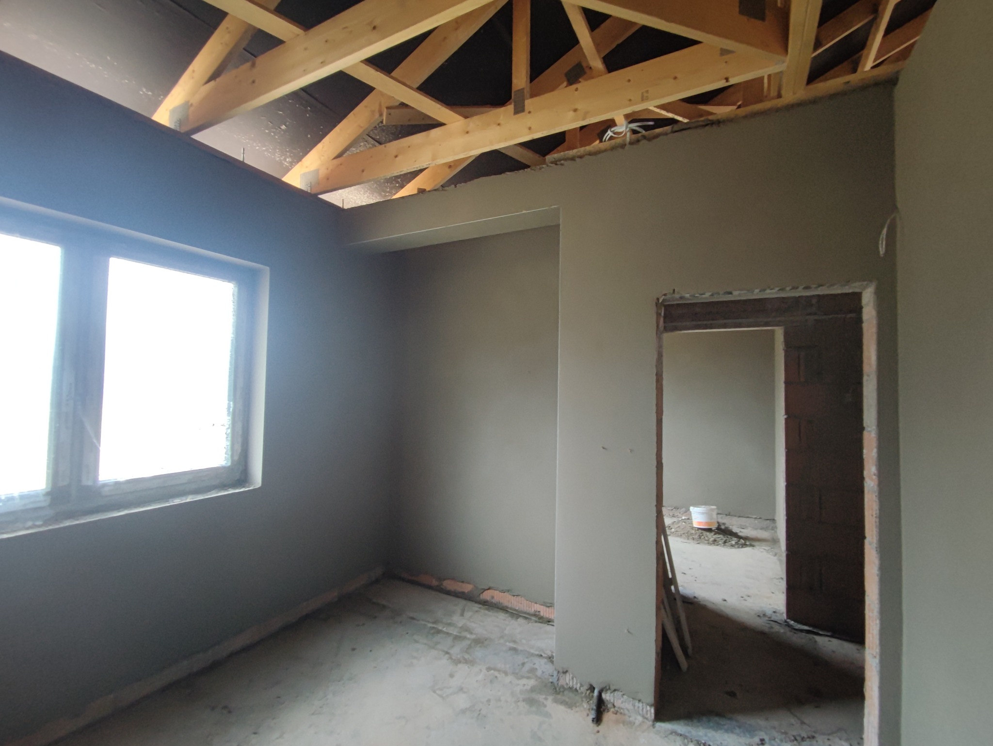 Wnętrze pomieszczenia w trakcie prac wykończeniowych: ściany pokryte świeżym, szarym tynkiem, widoczna drewniana konstrukcja dachu, surowa rama okienna i przejście do kolejnego pomieszczenia...