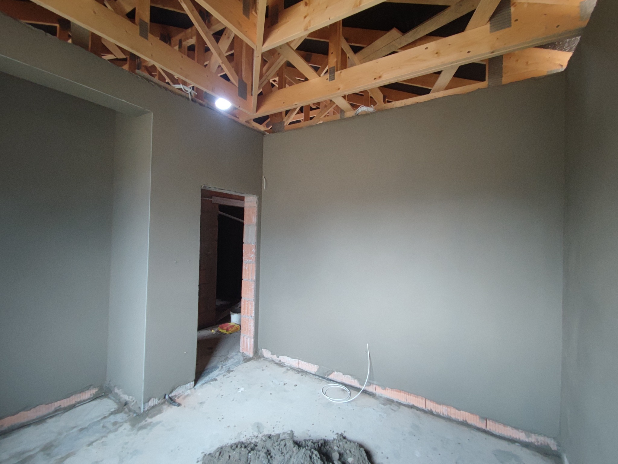 Wnętrze pomieszczenia w trakcie prac wykończeniowych: ściany pokryte świeżym, szarym tynkiem, widoczna konstrukcja drewniana dachu, kabel elektryczny zwisający ze ściany, resztki zaprawy...