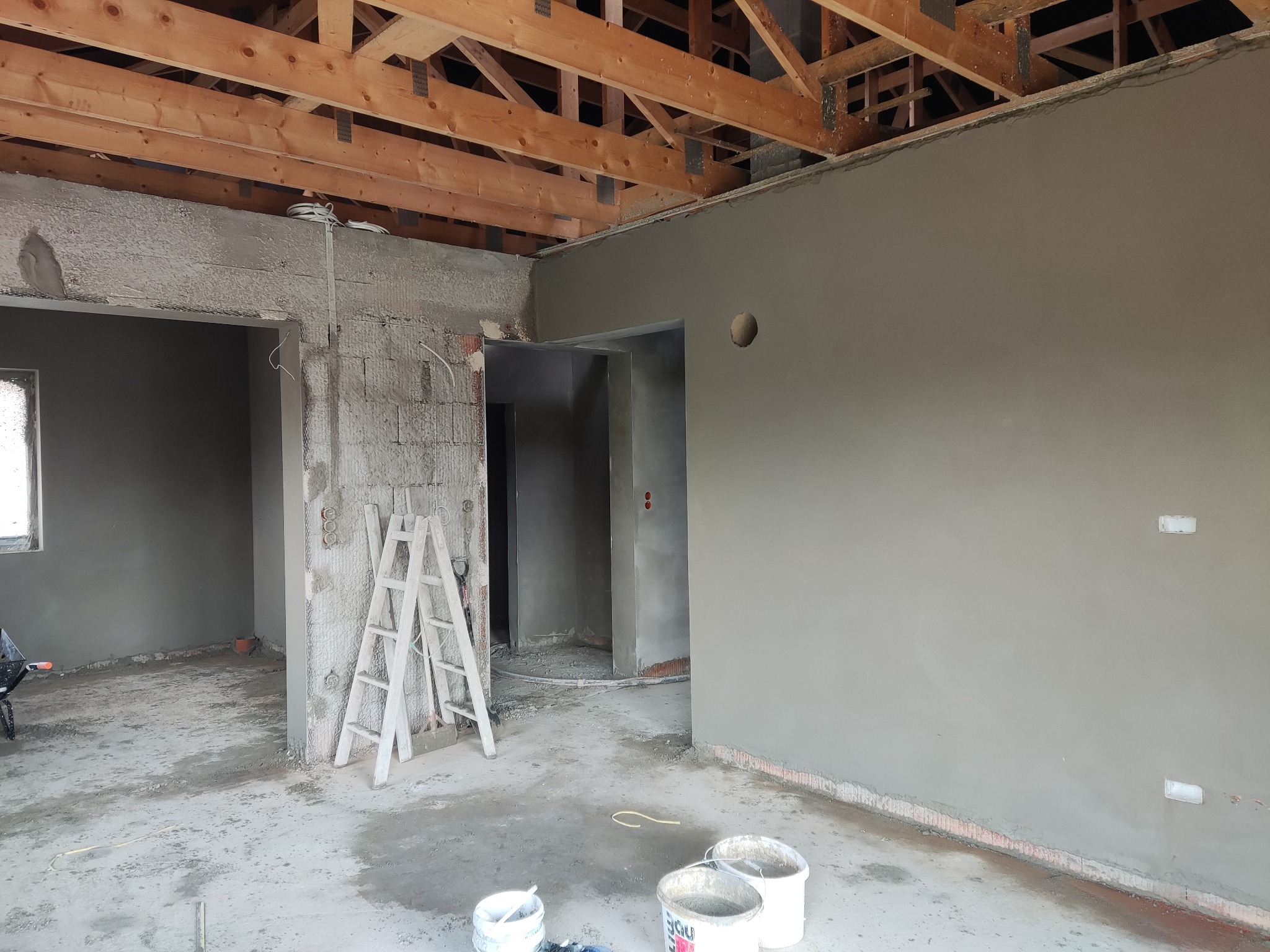Wnętrze domu w trakcie prac wykończeniowych: ściany pokryte świeżym, szarym tynkiem, widoczne drewniane elementy konstrukcji dachu, drabina oparta o ścianę z instalacją elektryczną, na podłodze...