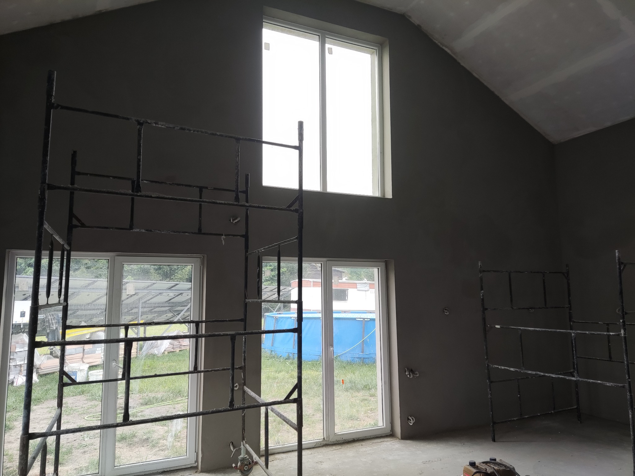 Wnętrze domu w trakcie tynkowania maszynowego, widoczne rusztowania, okna z widokiem na zewnątrz i surowa, szara powierzchnia ścian.