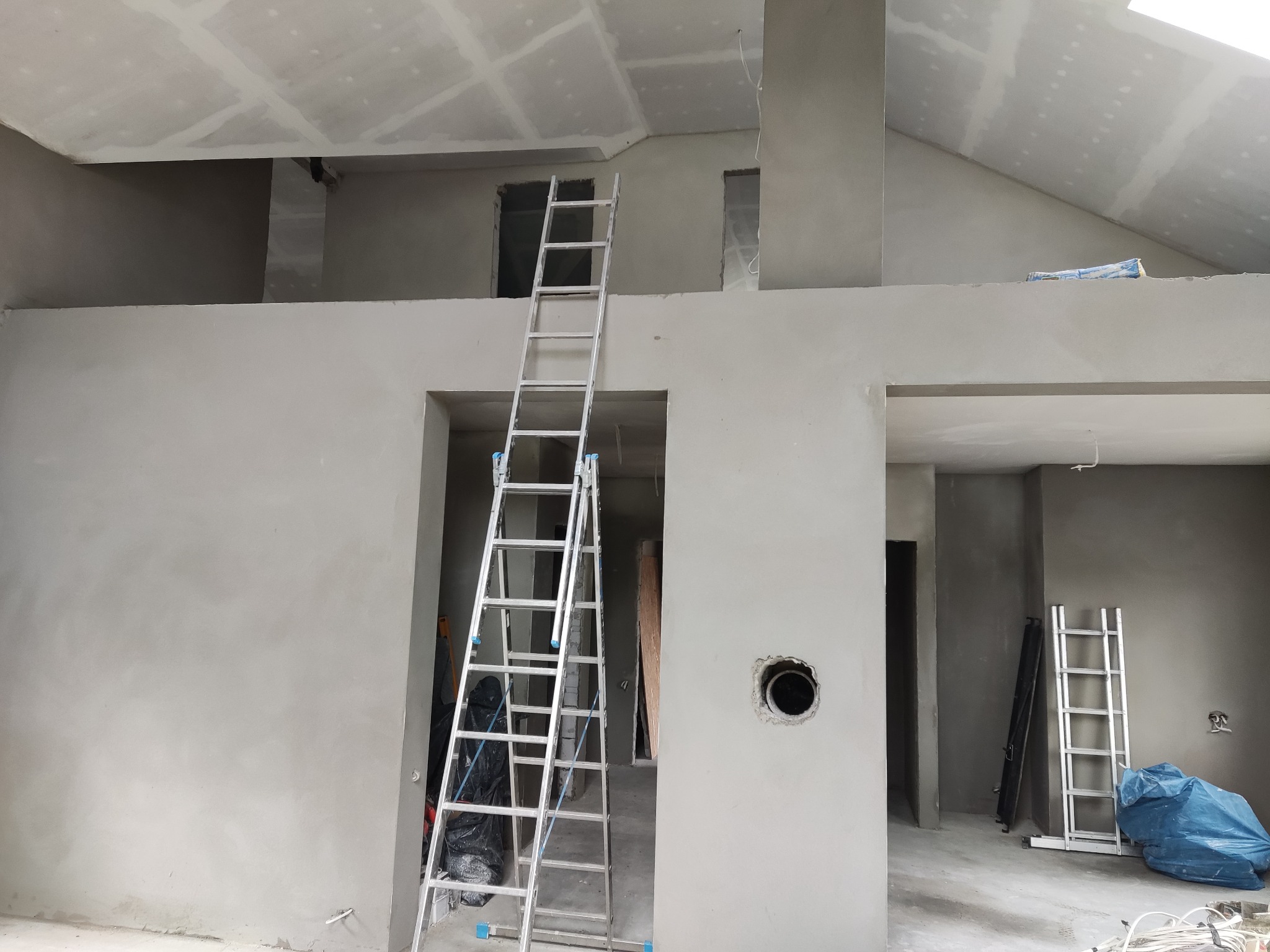 Wnętrze domu w trakcie prac wykończeniowych: ściany i sufit pokryte świeżym, jasnoszarym tynkiem, dwie aluminiowe drabiny rozstawione w pomieszczeniu, widoczny otwór instalacyjny w ścianie...