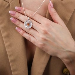La Crea Monika Nawrot - Kobieta w beżowym żakiecie prezentuje srebrny naszyjnik z ażurowym wzorem i bursztynowym oczkiem, dłoń z francuskim manicure na dekolcie.