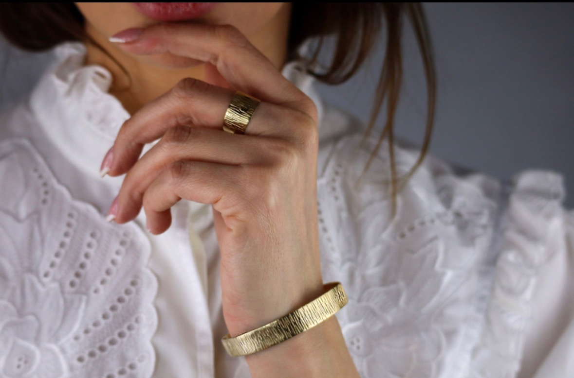 Kobieta prezentuje złotą biżuterię: szeroki pierścionek z fakturą kory drzewa na palcu i bransoletkę o podobnej fakturze na nadgarstku, na tle białej bluzki z haftem.