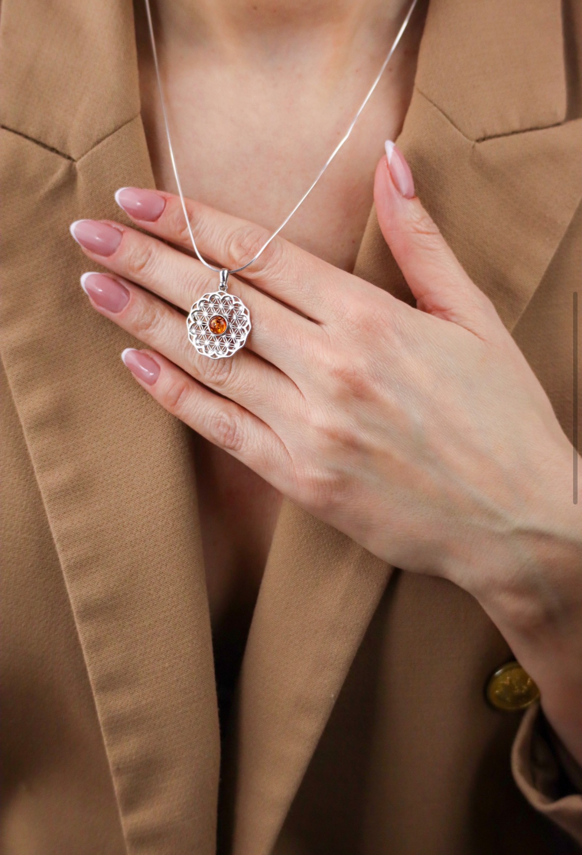 Kobieta w beżowym żakiecie prezentuje srebrny naszyjnik z ażurowym wzorem i bursztynowym oczkiem, dłoń z francuskim manicure na dekolcie.