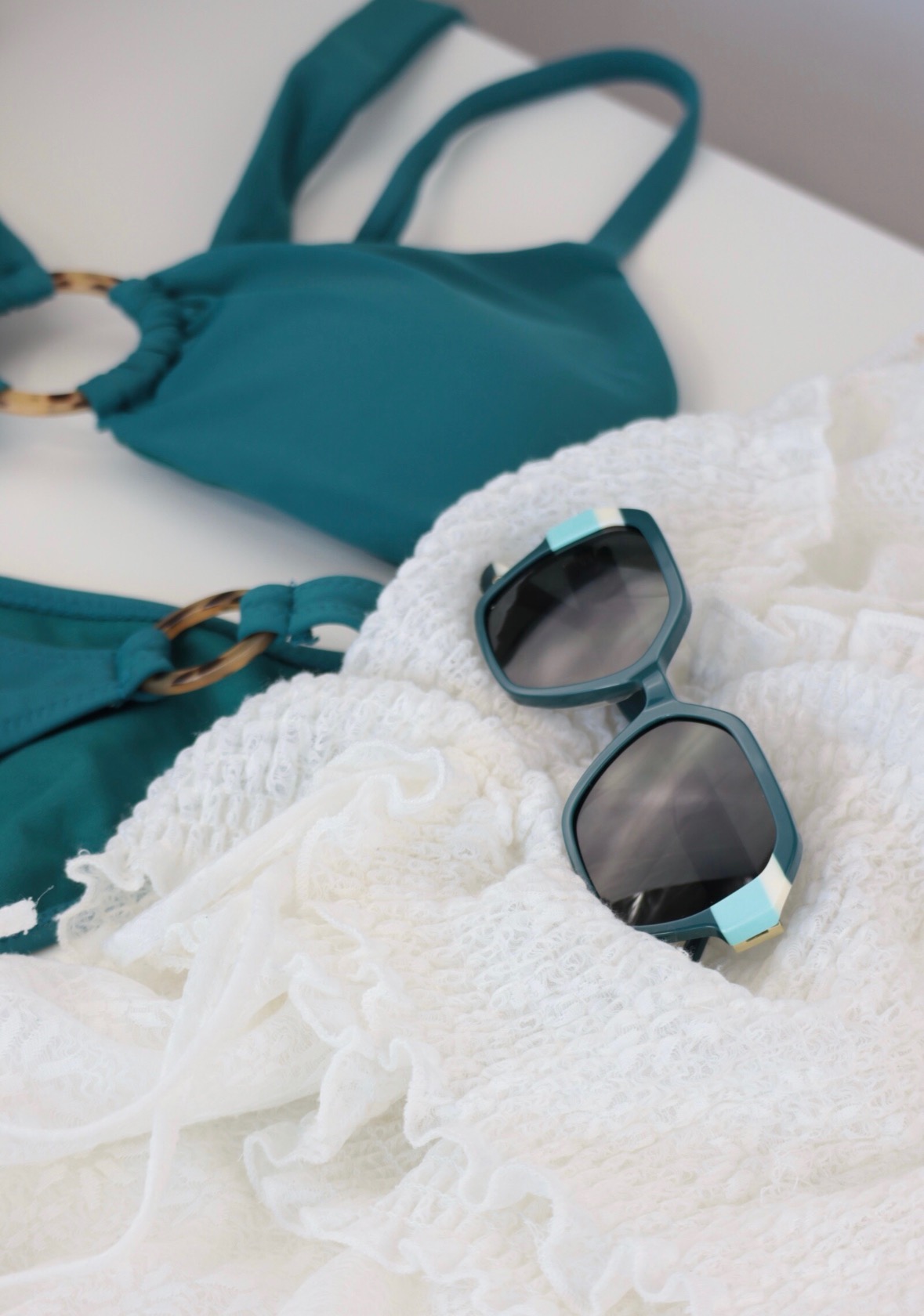 Turkusowy strój kąpielowy z okrągłymi detalami, biała ażurowa narzutka i turkusowe okulary przeciwsłoneczne z akcentami na białym tle.