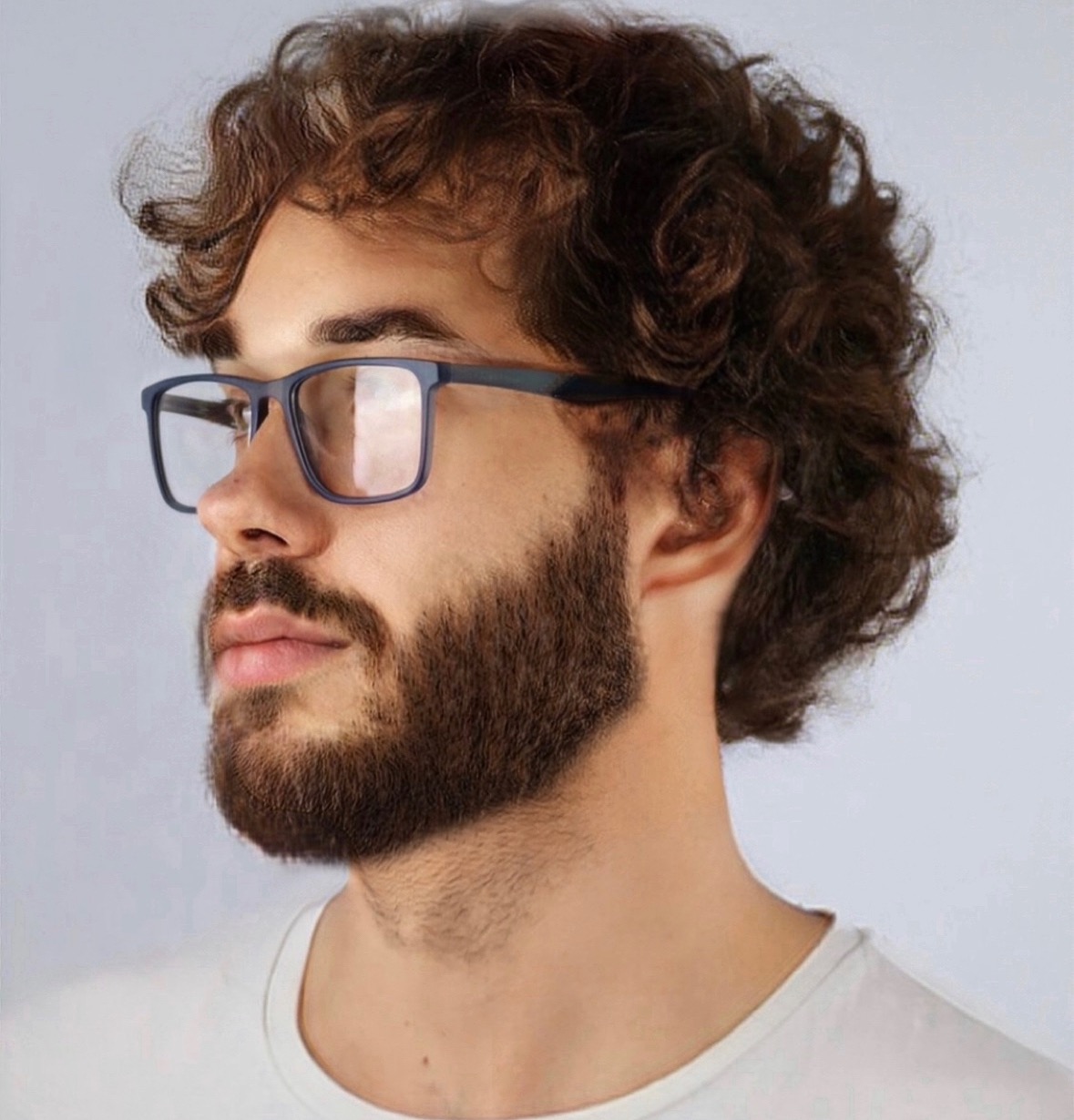 Portret mężczyzny z kręconymi włosami i brodą, w okularach z ciemnymi oprawkami, patrzy w lewo.