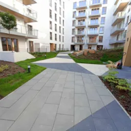 Nowoczesna ścieżka z szarych i grafitowych kostek brukowych prowadząca między budynkami apartamentowymi z zielonymi rabatami po bokach.