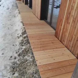 Jasna, drewniana ścieżka z desek prowadząca wzdłuż ściany budynku obłożonej drewnem, obok widoczne kamienie i resztki śniegu.